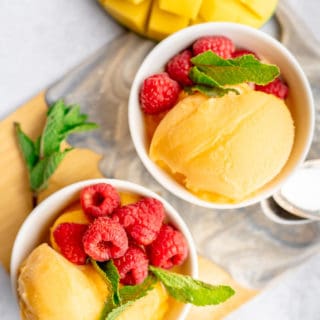 Two white bowls with scoops of mango sorbet, mint and fresh raspberries and a cut mango on the side
