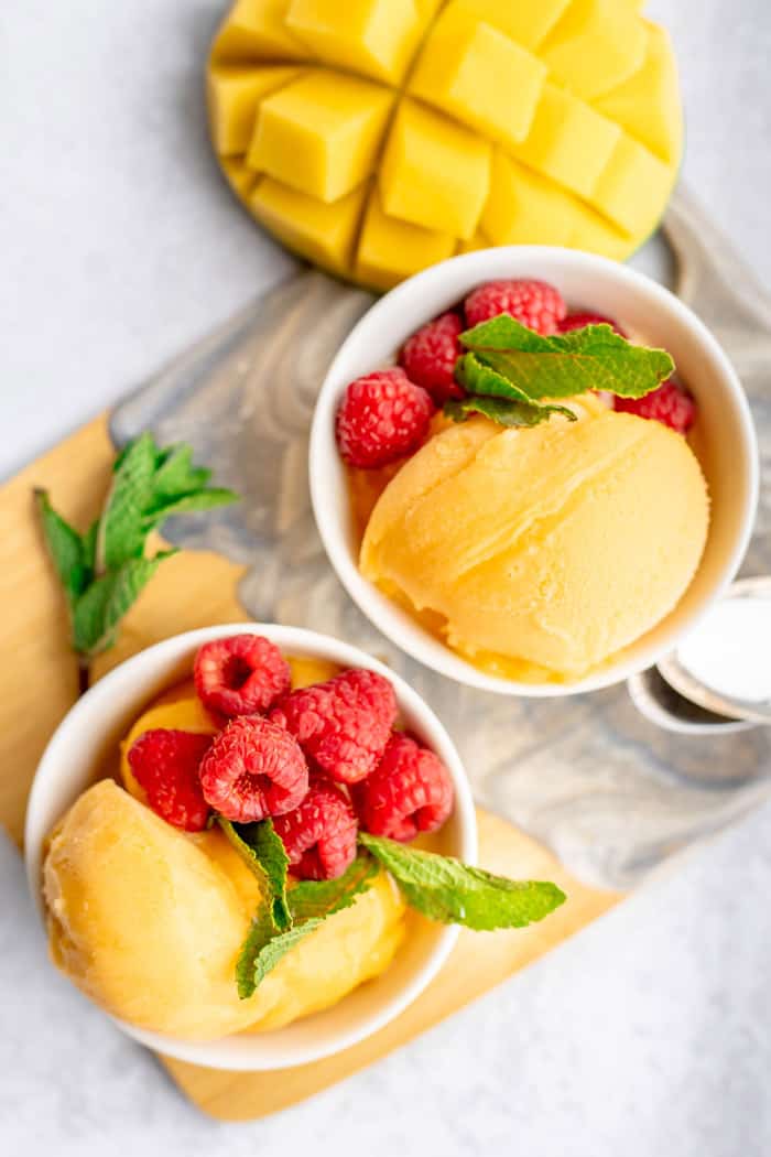 Two white bowls with scoops of mango sorbet, mint and fresh raspberries and a cut mango on the side