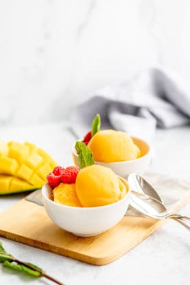 Two white bowls filled with mango sorbet, raspberries and fresh mint ready to serve against a white background