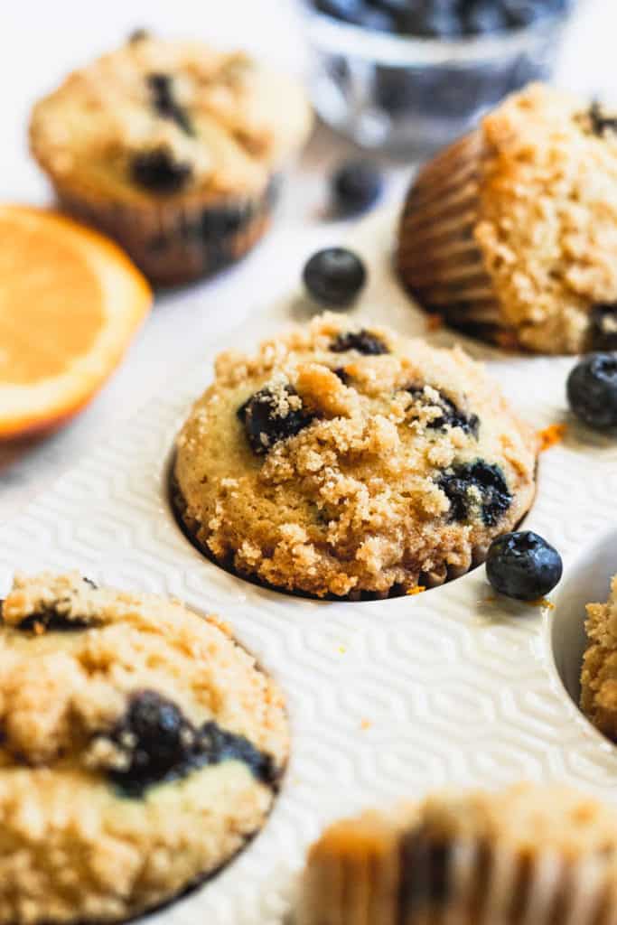A close up of blueberry muffins in tin after baking with orange slices and blueberries scattered around