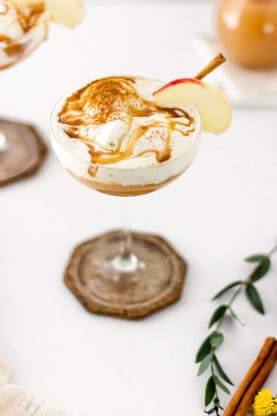 A glass of apple cider along with melted ice cream with a slice of apple and cinnamon stick on the side against white background