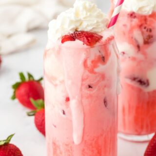 Glasses of strawberry floats on the table with a red and white straw and topped with berries and whipped cream.