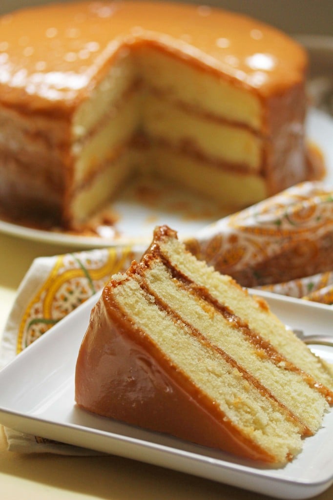 A slice of the 3-layer caramel cake on a white plate in the foreground with the caramel cake with a slice missing in the background.