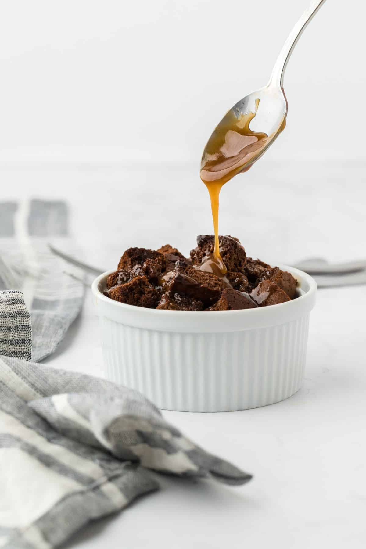 A ramekin of gingerbread bread pudding with a spoon of caramel sauce over it drizzling down.