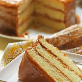 Slice of southern caramel cake on a plate the whole cake is in the background.