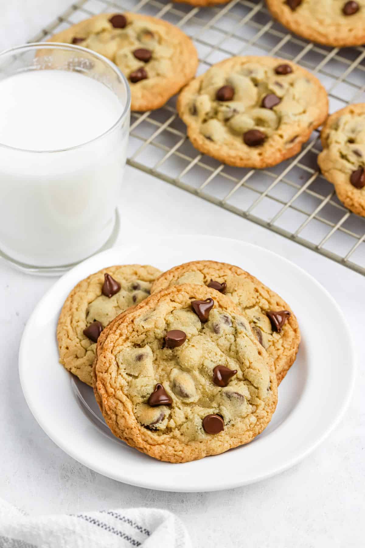 A plate of Mrs Fields cookies on a plate with more on a cooling rack in the back and a glass of milk.