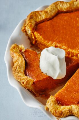 Two slices of sweet potato pie with a dollop of whipped cream on top sitting on their own white plates with forks next to the whole pie with the two cut out slices. 