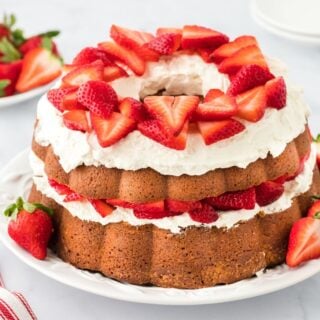 Strawberry shortcake pound cake with fluffy whipped cream and sliced strawberries topping each layer, displayed on a white plate with additional strawberries placed around it, all set in a marble surface with a striped red and white towel to the side and more strawberries in the background