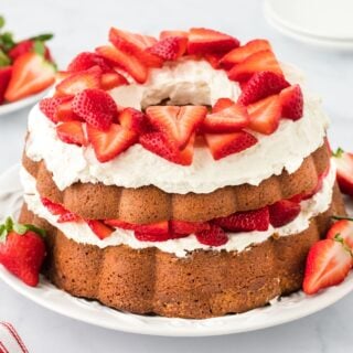 Strawberry shortcake pound cake with fluffy whipped cream and sliced strawberries topping each layer, displayed on a white plate with additional strawberries placed around it