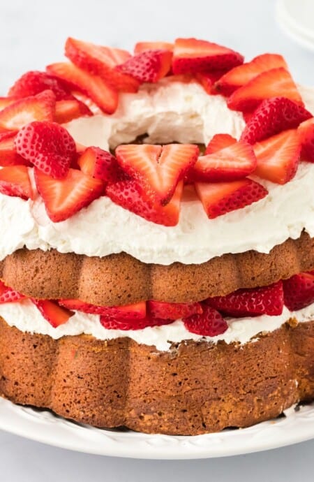 Strawberry shortcake pound cake with fluffy whipped cream and sliced strawberries topping each layer, displayed on a white plate with additional strawberries placed around it