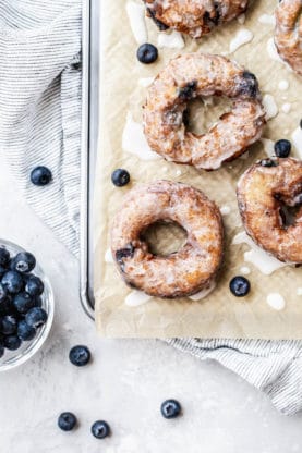 Overhead shot of Blueberry Glazed Doughnuts served on a parchment paper on a metal baking sheet with a bowl of blueberries next to them