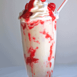 White chocolate raspberry milkshake contained in a clear glass topped with whipped cream and fresh raspberries and a straw.