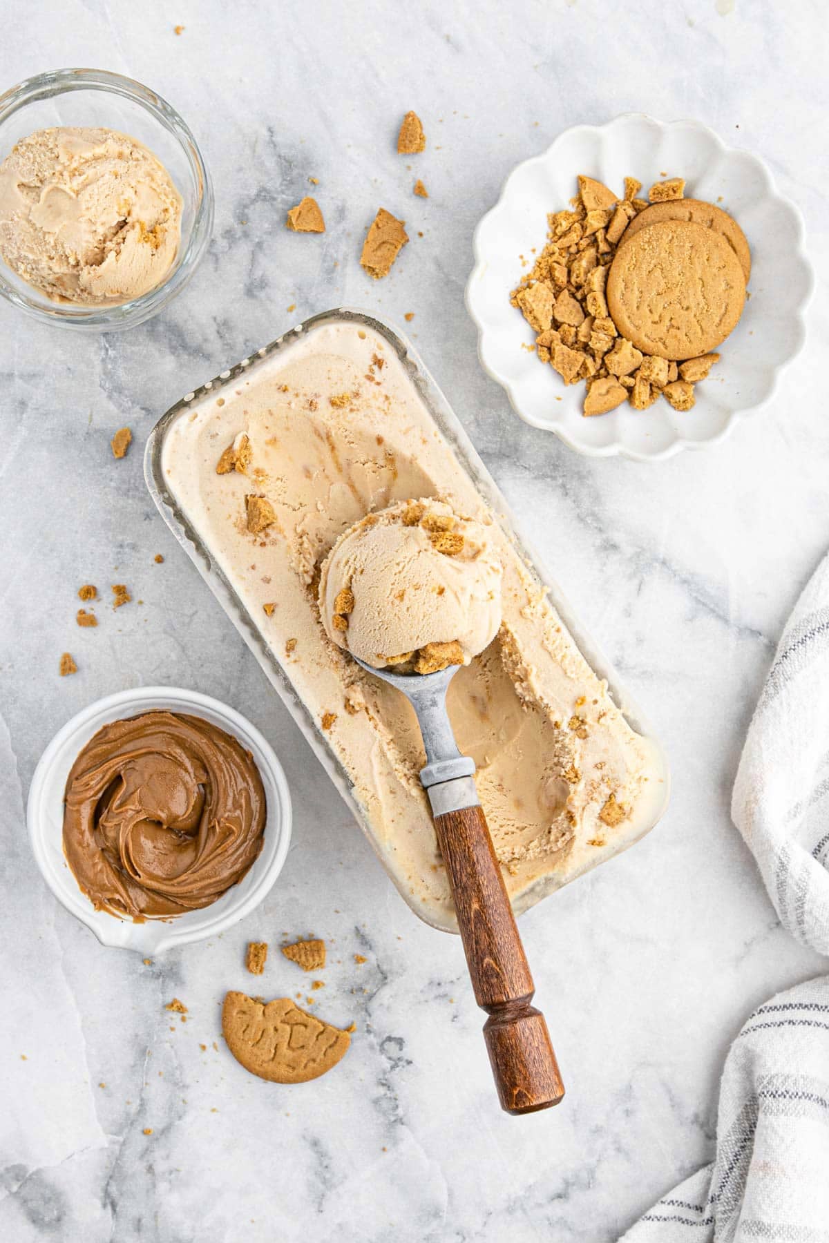 Overhead view of the table with cookie butter ice cream in a rectangle container with a scooper filled with ice cream on top and ingredients in bowls around it.