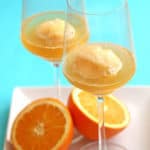 Orange sorbet mimosas contained in two wine glasses and resting on a square, white plate with an orange sliced in half between the glasses