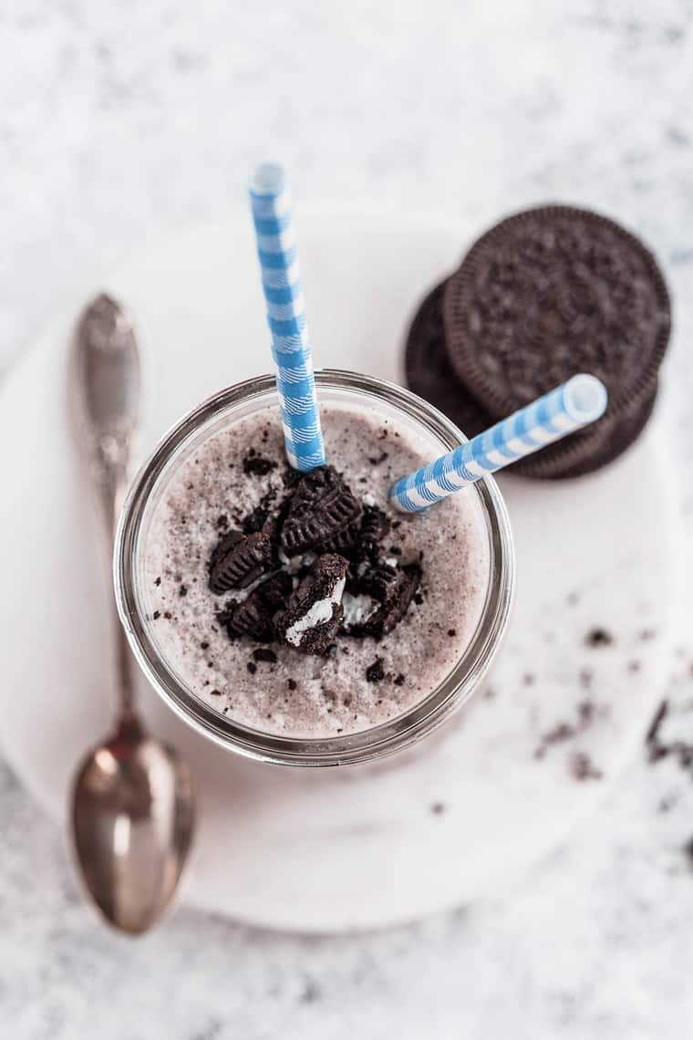 Overhead shot of a glass of oreo milkshake with two blue and white striped straws and a few oreo cookies stacked on top of each other and a spoon next to it