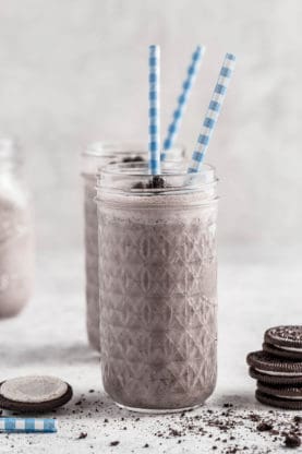 Two light oreo smoothies with two blue and white striped straws and a few oreo cookies stacked on top of each other next to it