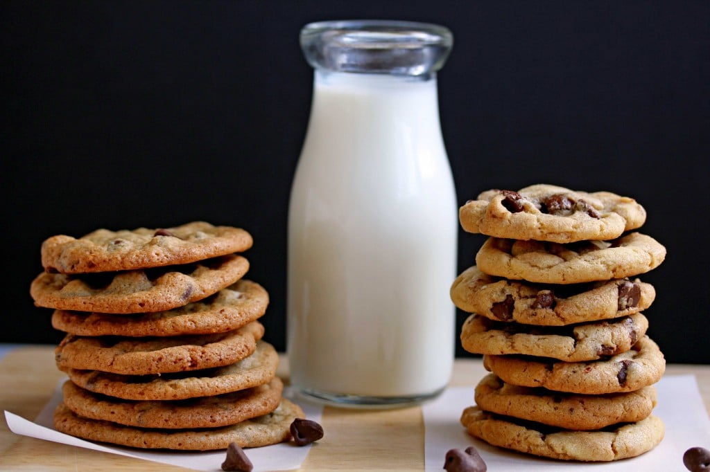 A stack of crispy chocolate chip cookies on the left and a stack of chewy chocolate chip cookies on the right with a glass jug of milk in between the two stacks