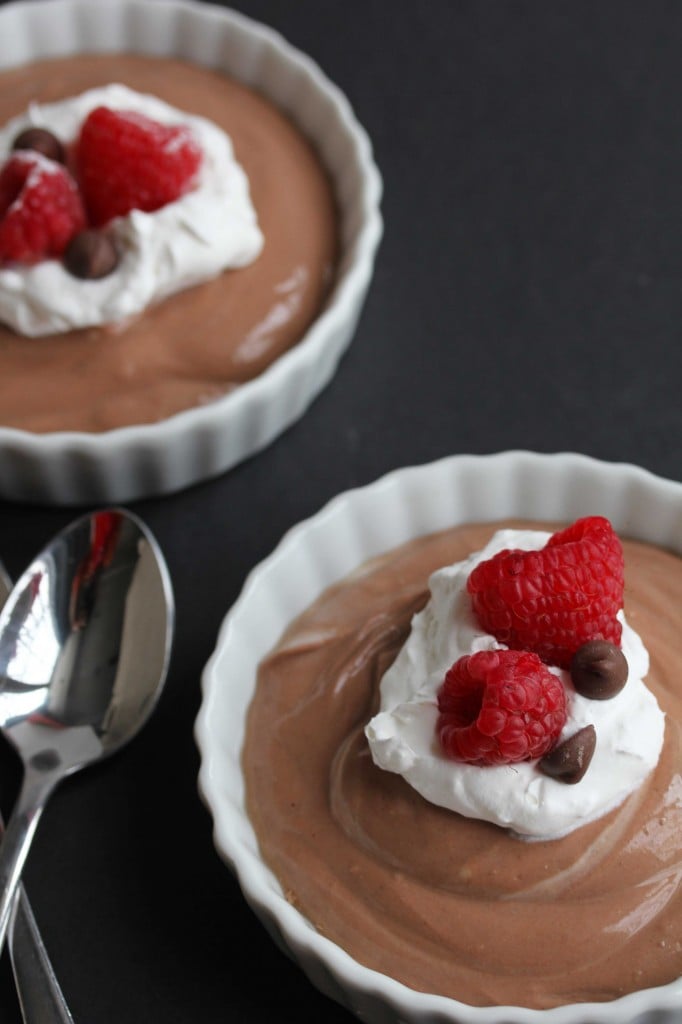 Light chocolate mousse contained in two circular, white bowls topped with whipped cream, raspberries and chocolate chips with two silver spoons