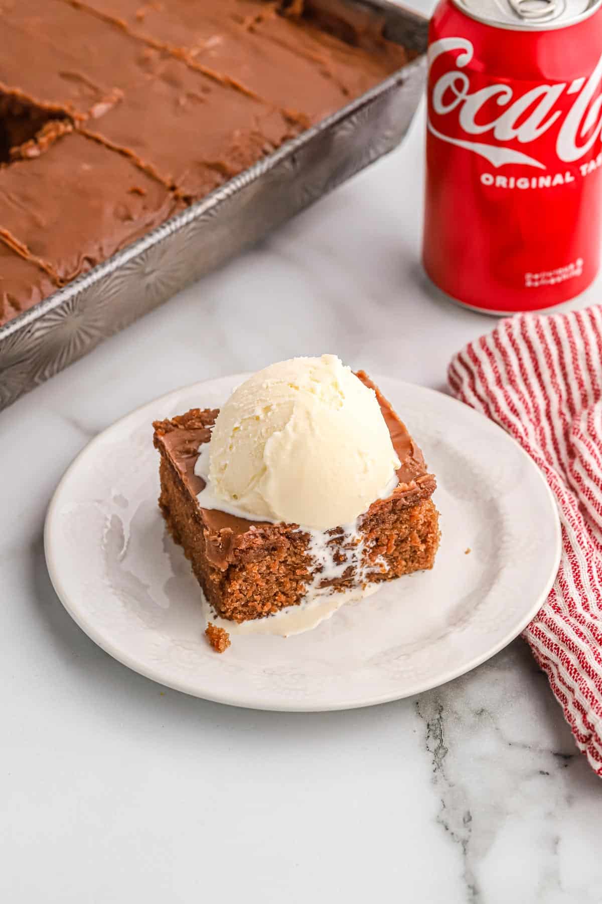 Coca cola cake slice on a white plate with a scoop of vanilla ice cream on top and a can of coca cola behind.