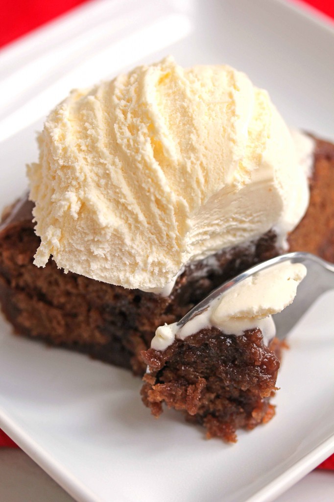 A close up of a slice of warm coca cola chocolate cake topped with a scoop of vanilla ice cream and a spoon going through it