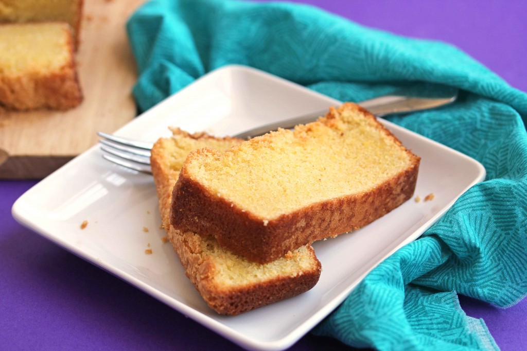 Two slices of cornmeal pound cake stacked on top of one another on a square, white plate with a fork on a purple background and a greenish cloth