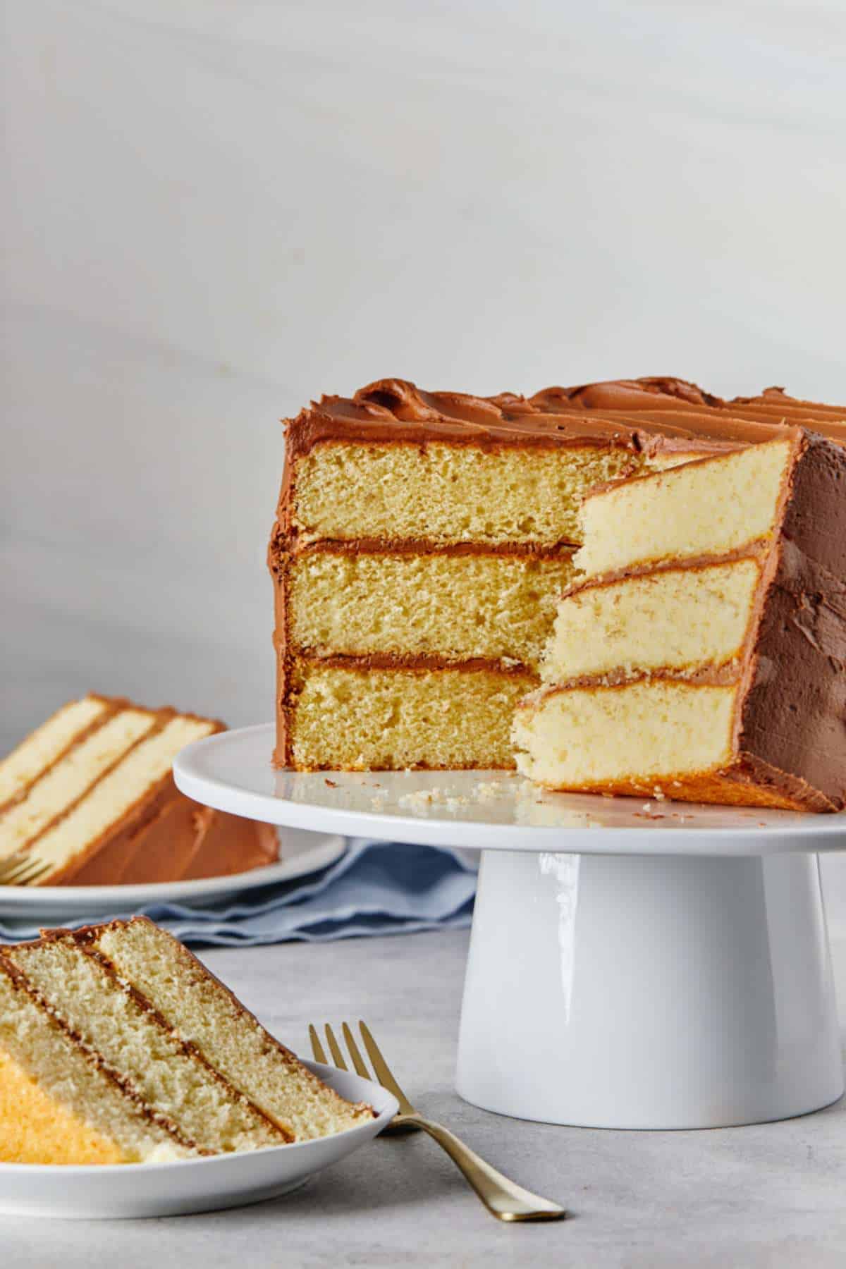 A yellow cake with chocolate frosting on a white cake stand with pieces missing and a slice on a plate in the front with a fork beside the plate.