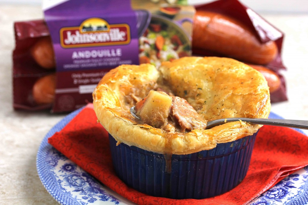 Creole pot pie in a small blue bowl with a spoon in it holding sausage and a piece of potato with an unopened package of andouille sausage in the background