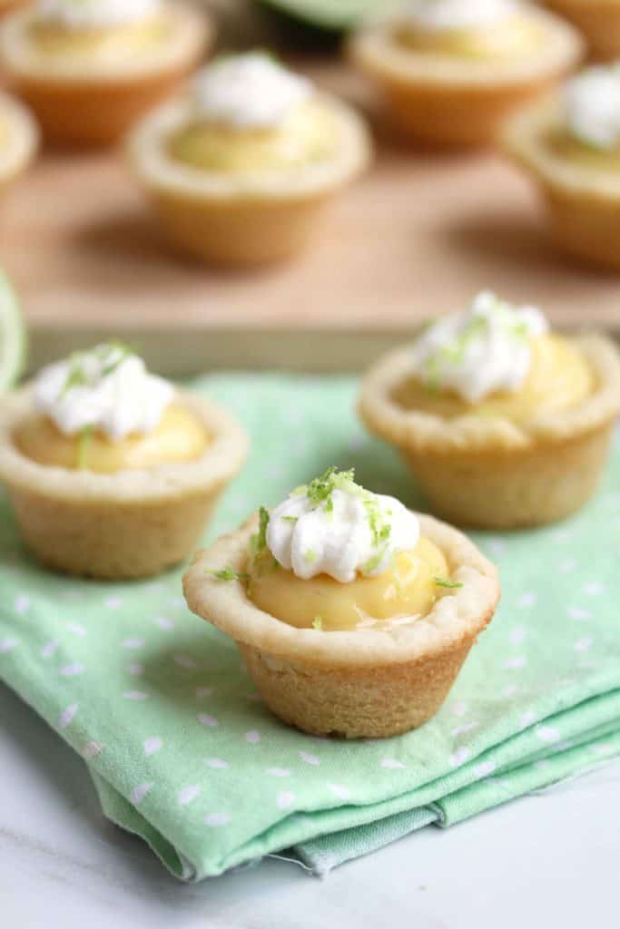 Three key lime pie cookie cups sitting on a light green cloth and several more in the background