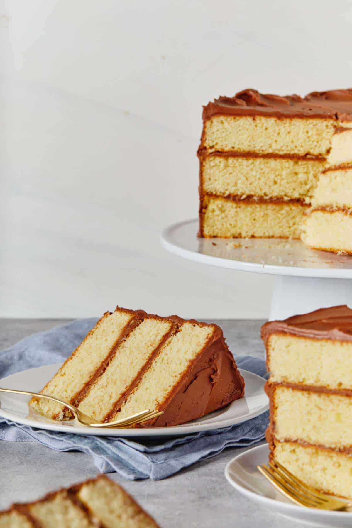 Yellow butter cake with chocolate frosting on white plates with some cake on a stand in the background.