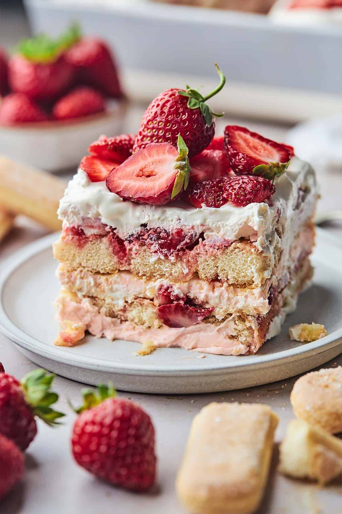 A square of strawberry tiramisu on a plate topped with a whole strawberry and some sliced.