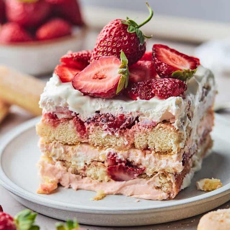 Close up of a piece of strawberry shortcake tiramisu sitting on a square, white plate with a bowl of strawberries in the background