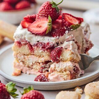 Strawberry Shortcake Tiramisu slice on a white plate surrounded by strawberries and lady fingers in background