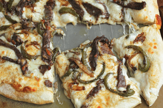 Close up of one slice from this Cheese Steak Pizza being pulled from the pie with cheese clinging to the slice and the pizza pie.