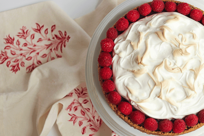 Overhead shot of a raspberry s'more pie displayed on a white cake stand with white and red linen underneath it