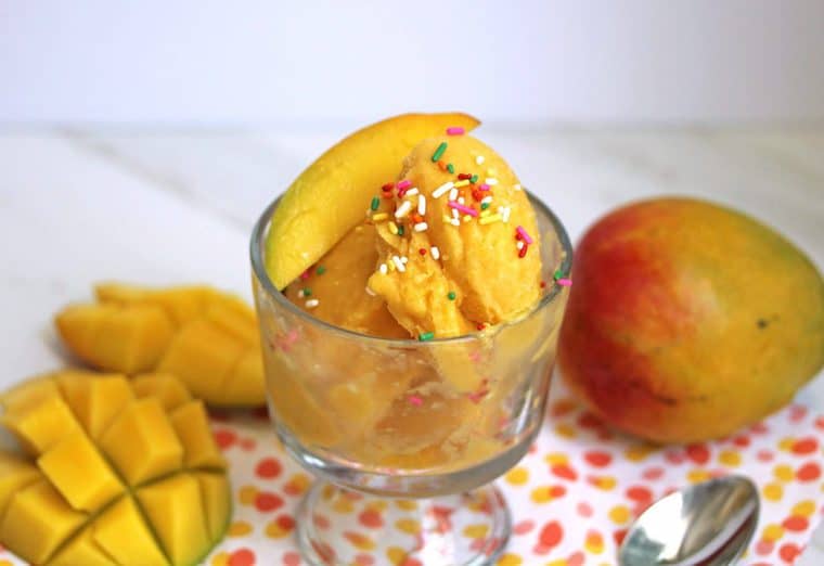 A few scoops of mango frozen yogurt in a glass bowl topped with sprinkles and a slice of mango and fresh mangoes around the bowl as decoration