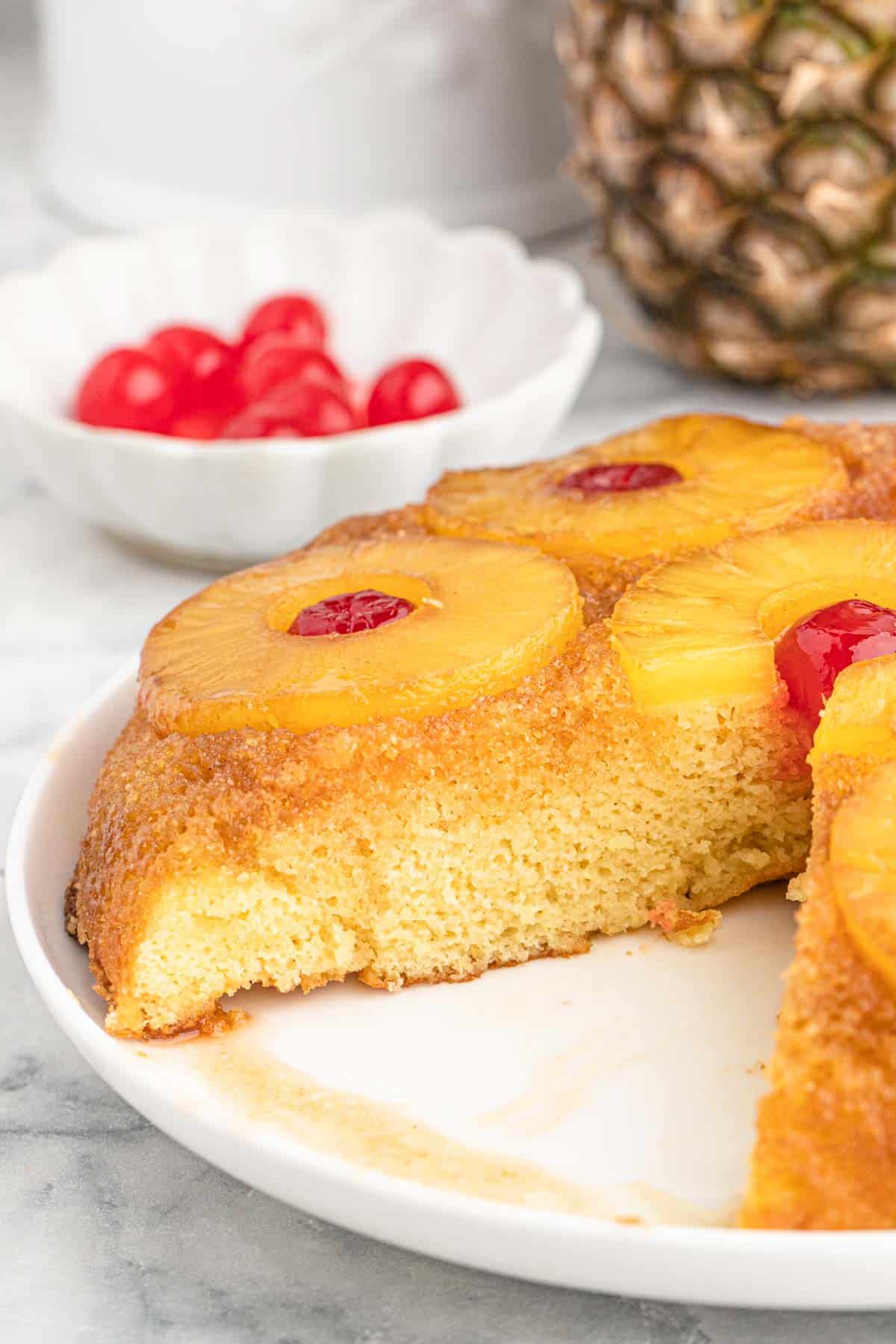 A pineapple upside down cake on a white platter with a slice cut out and a bowl of cherries in the background.