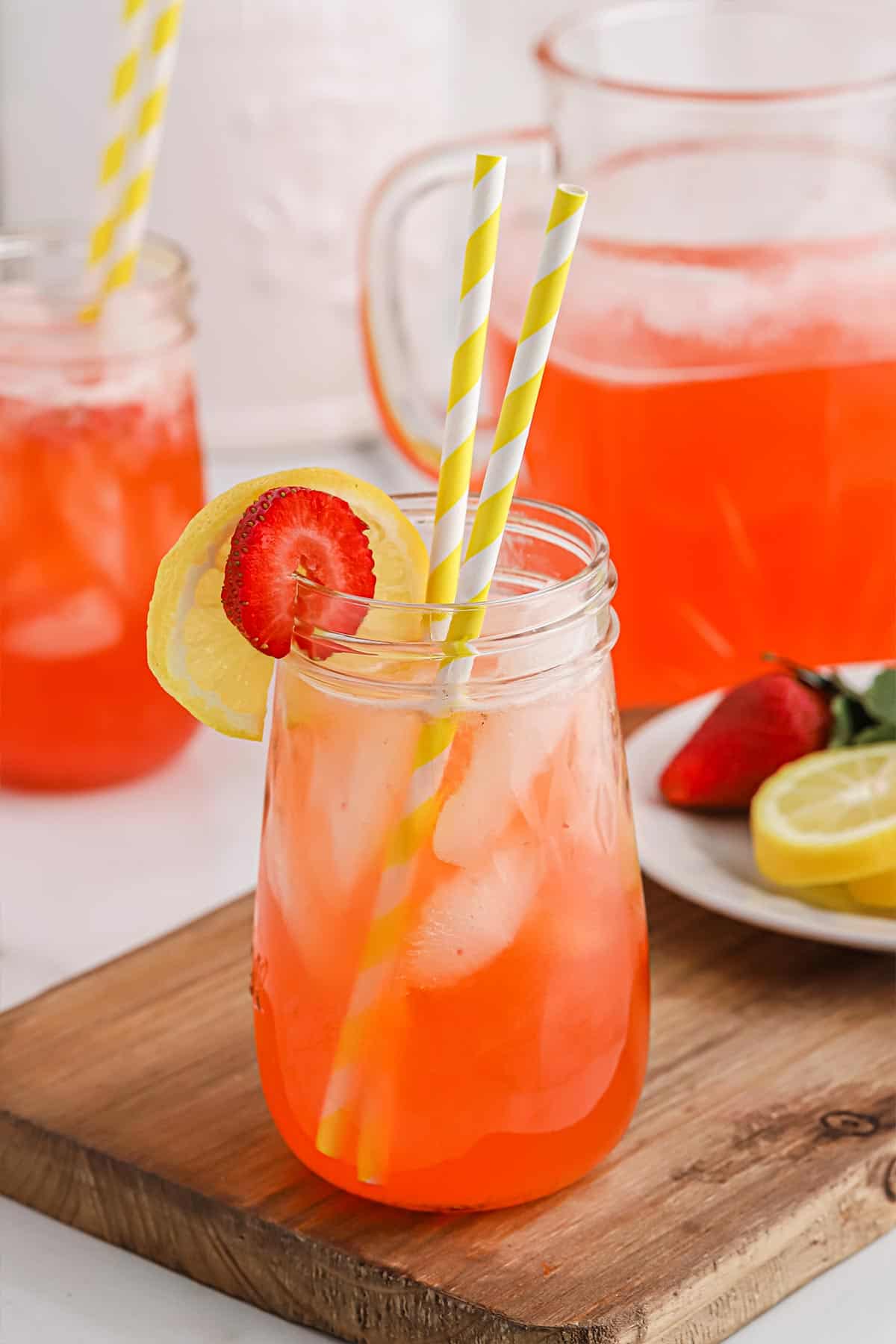 A glass of strawberry lemonade poured up with the pitcher behind and garnished with lemon slice and strawberry.