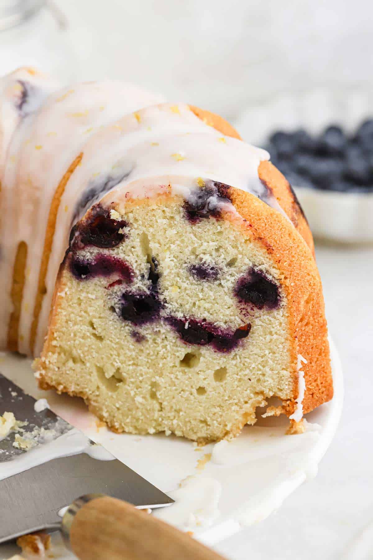 Lemon blueberry cake cut to show the inside studded with blueberries and a glaze and lemon zest on top.