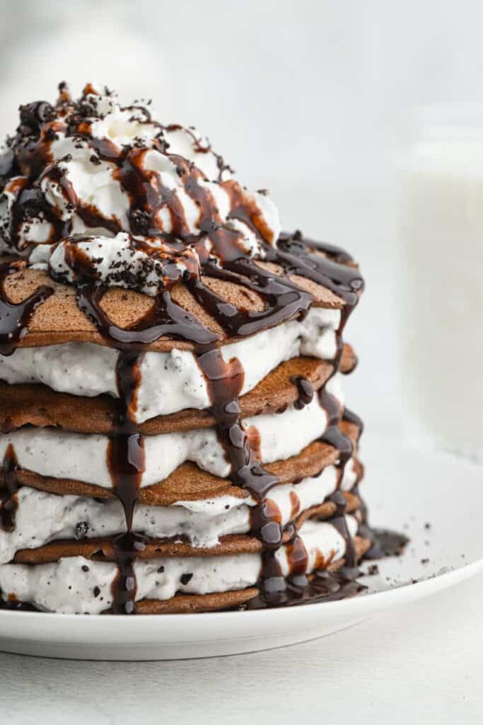 Stack of oreo pancakes topped with crumbled oreo cookies, whipped cream and chocolate sauce on a white plate with a fork