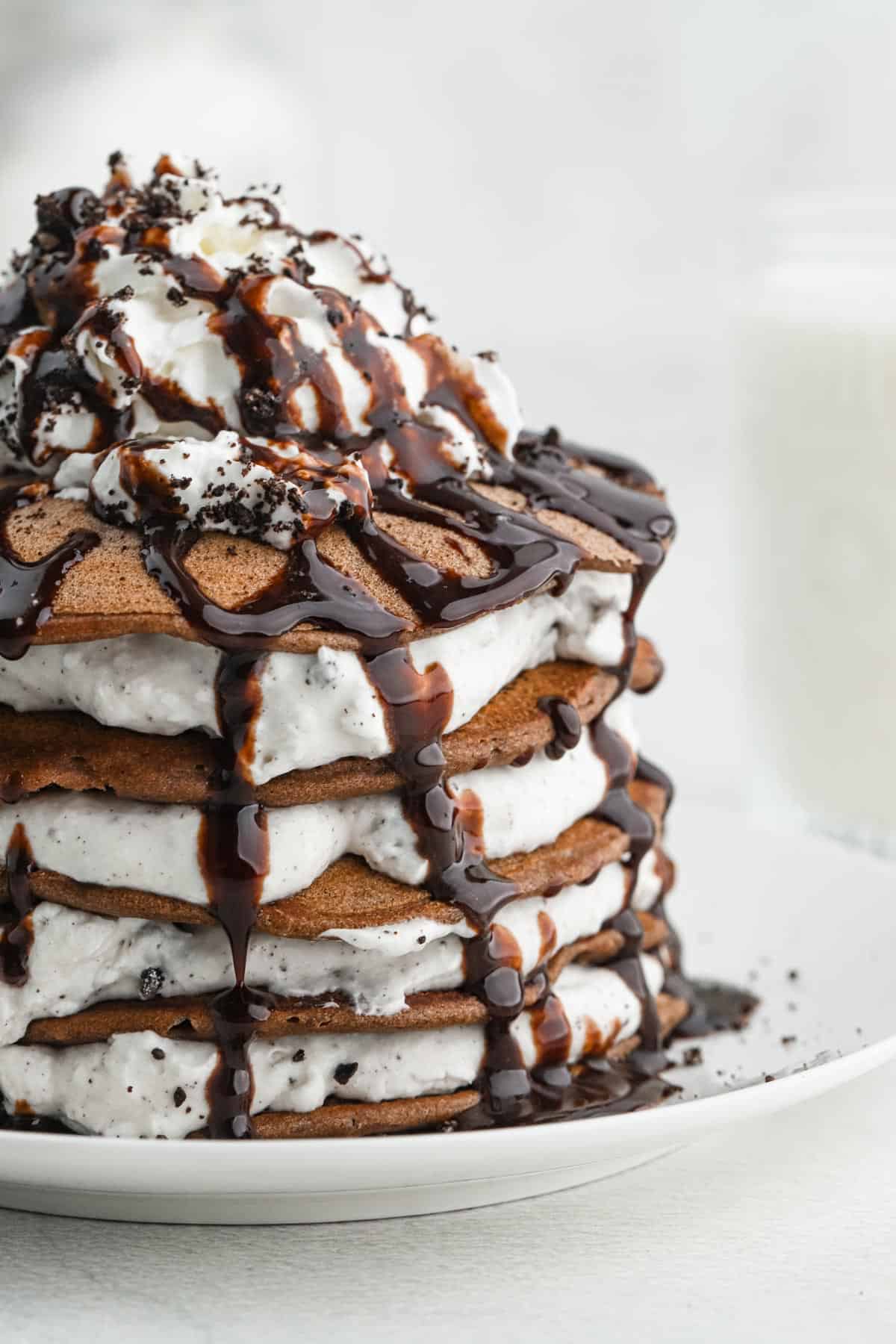 Stack of oreo pancakes topped with crumbled cookies and chocolate sauce, layered with whipped cream on a white plate.