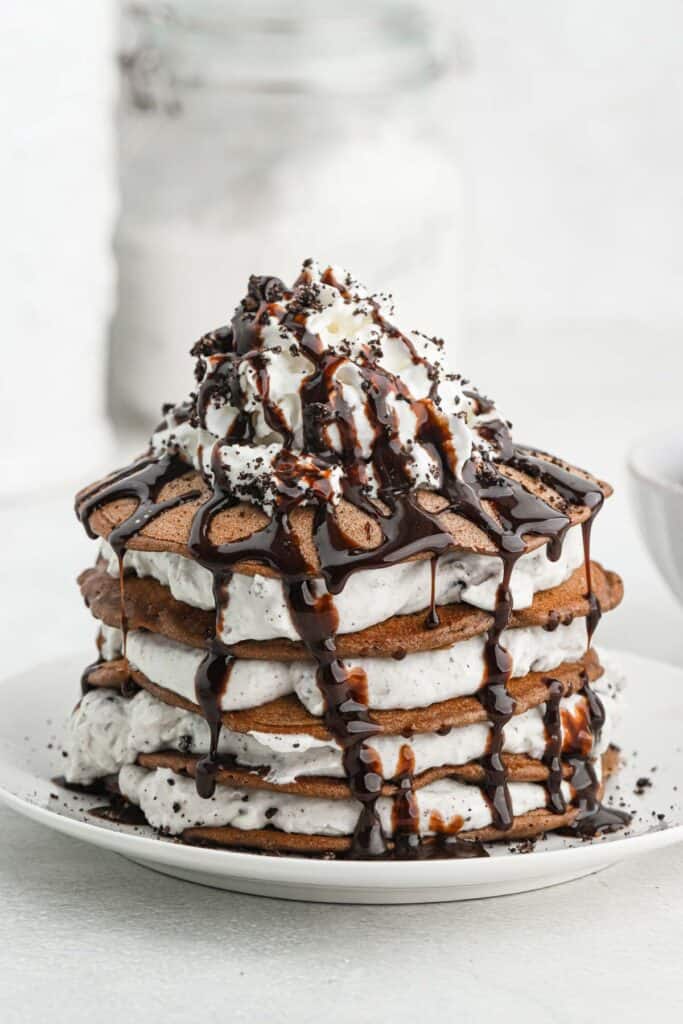 From above, a stack of oreo pancakes topped with crumbled oreo cookies, whipped cream and chocolate sauce on a white plate with a piece taken out