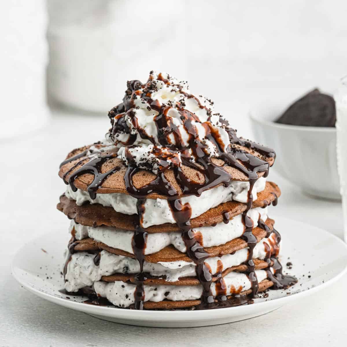 Close up of a stack of oreo pancakes topped with crumbled oreo cookies, whipped cream and chocolate sauce on a white plate with a piece taken out