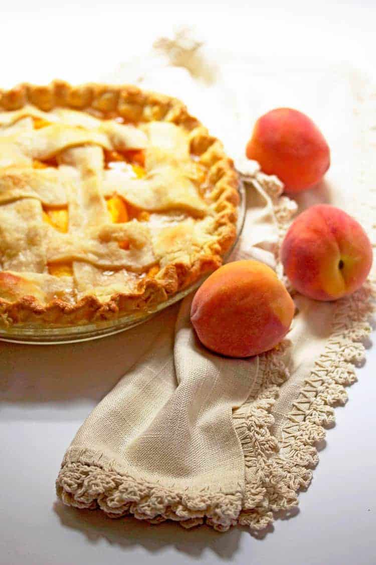 A peach pie with lattice crust on a white background with scattered fresh peaches against beige napkin.
