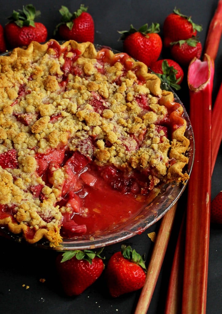 Close up of a strawberry rhubarb crumble pie with fresh strawberries and rhubarb next to it and a slice missing