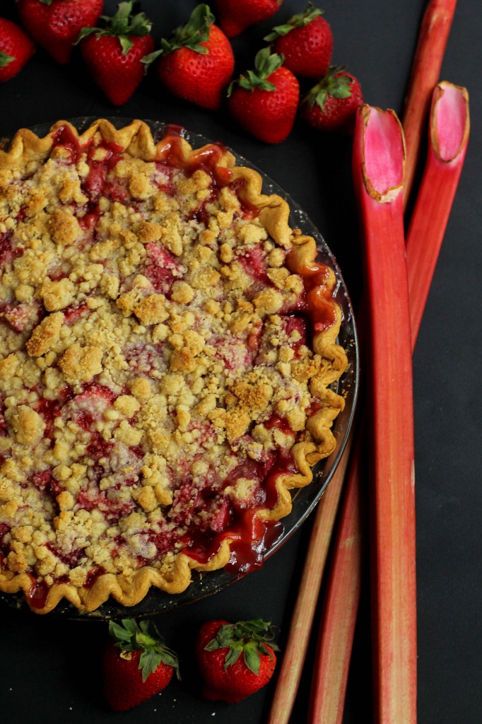 Overhead shot of a strawberry rhubarb crumble pie with fresh strawberries and rhubarb next to it
