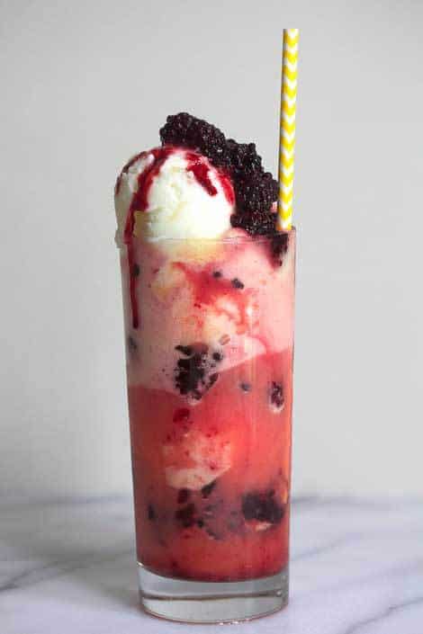 A blackberry pineapple float contained in a clear glass with a white and yellow striped straw in it