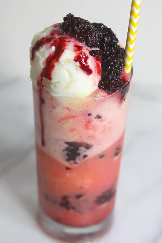 From above, a blackberry pineapple float contained in a clear glass with a white and yellow striped straw in it