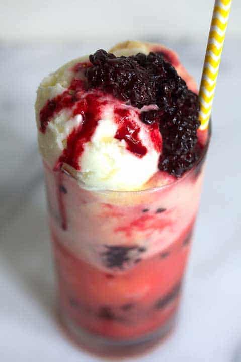 Close up of a blackberry pineapple float contained in a clear glass with a white and yellow striped straw in it