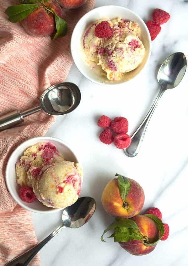 Overhead shot of two white bowls filled with peach raspberry ice cream decorated with an ice cream scoop, spoons, fresh peaches and raspberries