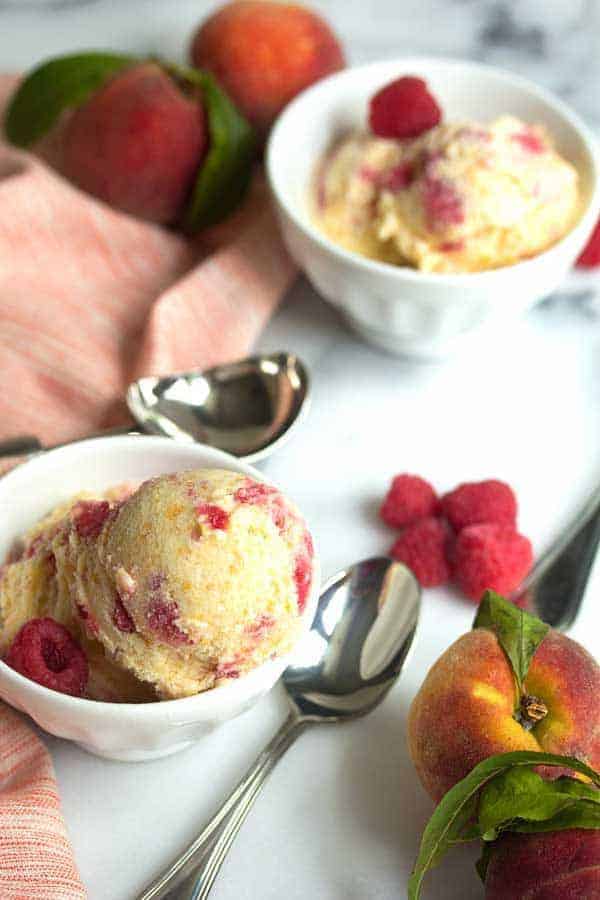 Two white bowls filled with peach raspberry ice cream decorated with an ice cream scoop, spoons, fresh peaches and raspberries
